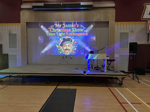 A school gym stage with a white piano and the Christmas show title on the big screen