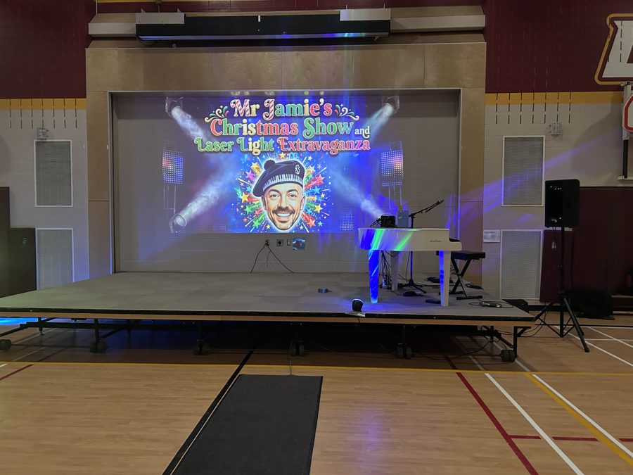 A school gym stage with a white piano and the Christmas show title on the big screen