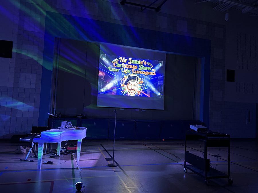 A school gym set for the Christmas Sing-Along: white piano under blue and green light, the show title on the big screen