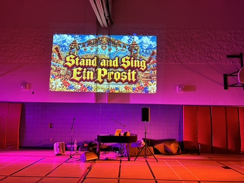 A school gym set for the Oktoberfest Assembly: the piano and sound system under purple stage light, with the big screen lit for the sing-along