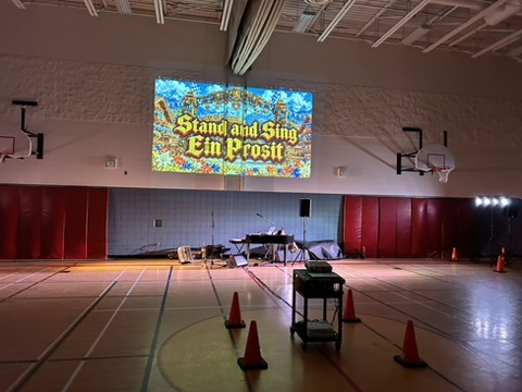 The same gym in daylight during setup: piano, sound system and screen at the stage end, floor space for the whole school in front